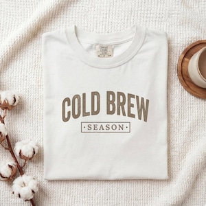 May include: A white short-sleeved t-shirt with a crew neckline featuring the brown text COLD BREW SEASON in a bold, arched font. The shirt has a Comfort Colors brand label at the inner neck area, showing the size S and the letters CM.