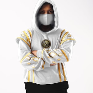 May include: A white hooded sweatshirt with gold trim and a gold circular design on the chest. The hood is pulled up and the person is wearing a white face mask.