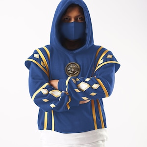May include: A blue hooded sweatshirt with gold trim and a wolf emblem on the chest. The hood is pulled up and the person is wearing a blue face mask.