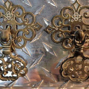 May include: Two ornate, antique brass drawer pulls with intricate floral and scrollwork designs. The pulls are mounted on a textured, diamond-plate metal surface, showcasing their aged patina and detailed craftsmanship. Ideal for vintage furniture restoration.