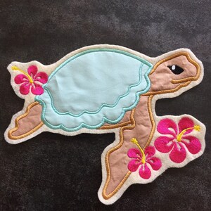 Könnte beinhalten: Eine Applikation aus hellblauem und braunem Filz, die eine Schildkröte mit rosa Hibiskusblüten zeigt. Die Schildkröte schwimmt mit erhobenem Kopf und ausgestreckten Flossen.