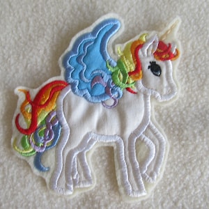 Könnte beinhalten: Ein weißes Filz-Einhorn mit Regenbogenmähne und -schwanz, blauen Flügeln und einem goldenen Horn. Das Einhorn steht auf einem weißen Hintergrund.