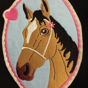 Könnte beinhalten: Gestickter Aufnäher mit dem Kopf eines braunen und weißen Pferdes mit schwarzer Mähne und einer rosa Blume in der Mähne. Das Pferd trägt ein weißes Zaumzeug. Der Aufnäher ist von einem weißen Oval mit rosa gewelltem Rand umgeben.