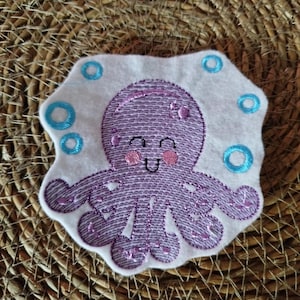 Könnte beinhalten: Ein lila Oktopus, der auf einem weißen Filzaufnäher gestickt ist. Der Oktopus hat ein lächelndes Gesicht und rosa Wangen. Blaue Kreise umgeben den Oktopus. Der Aufnäher befindet sich auf einer gewebten, braunen Oberfläche.