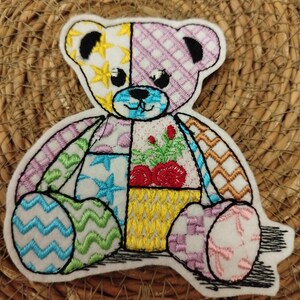 Könnte beinhalten: Gestickter Teddybär-Aufnäher mit Patchwork-Design. Der Bär hat Abschnitte mit gelben Sternen, rosa Karos, blauen Sternen, roten Blumen und grünen Zickzacklinien. Der Bär ist schwarz umrandet und sitzt auf einer gewebten Oberfläche.