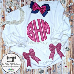 May include: White sweatshirt with a pink sparkly monogram "BHM" in a circle. The sweatshirt has two pink sparkly bows on the sleeves.