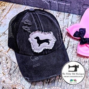 May include: Black distressed trucker hat with a black dachshund silhouette patch on a white background. The hat has a mesh back and a black brim.