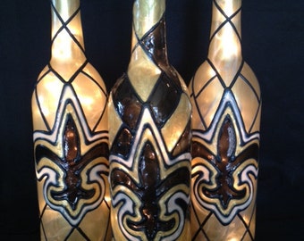 Puede incluir: Tres botellas de vino de vidrio decoradas con un diseño de flor de lis dorado, negro y blanco. Las botellas están iluminadas desde el interior, creando un brillo cálido.