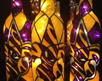 Puede incluir: Tres botellas de vino de vidrio decoradas con diseños de vitrales en amarillo, morado y negro. Las botellas están iluminadas desde adentro, creando un brillo cálido. Los diseños presentan un motivo de flor de lis.