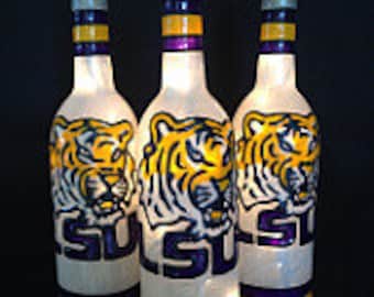 Puede incluir: Tres botellas de vino de vidrio transparente con fondo blanco y una franja púrpura y dorada alrededor de la parte superior. Cada botella tiene una imagen pintada de un tigre con las letras LSU en púrpura en la parte inferior.