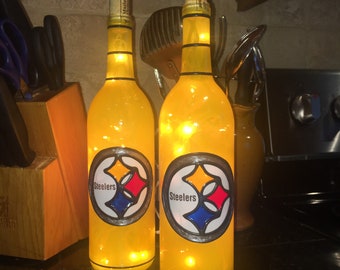 Puede incluir: Dos botellas de vino de vidrio amarillo con tapas de corcho y luces de cadena en el interior. Cada botella tiene un logotipo de Steelers pintado en el frente con las palabras "Steelers" en negro.