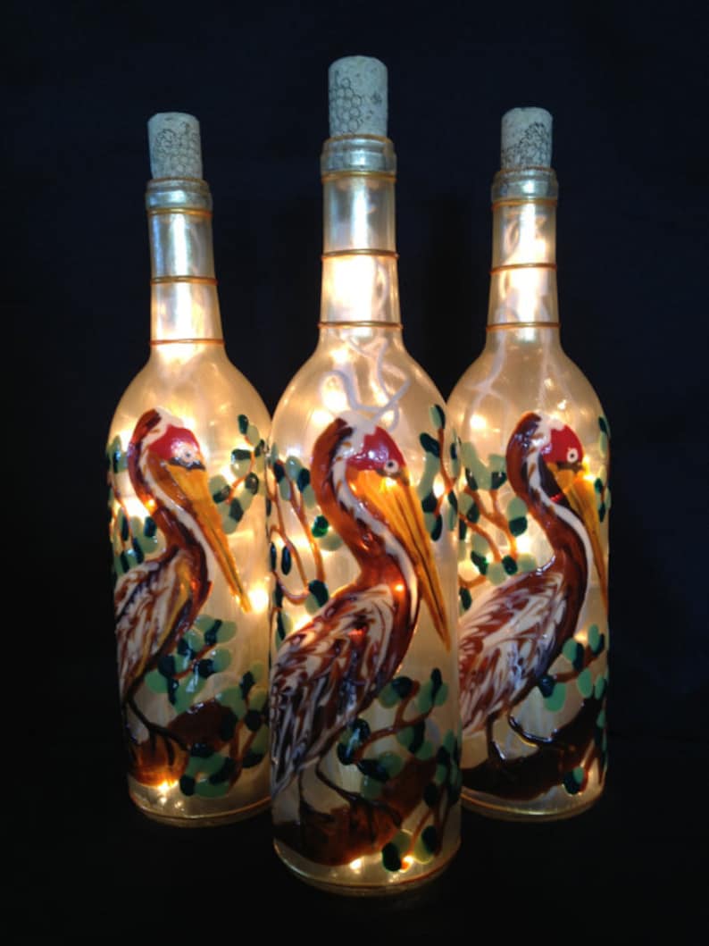 Puede incluir: Tres botellas de vidrio con tapas de corcho, cada una decorada con una imagen pintada de un p&aacute;jaro marr&oacute;n y blanco con un pico largo, de pie en una rama con hojas verdes. Las botellas est&aacute;n iluminadas desde adentro con luces blancas.