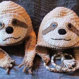 May include: Two handmade crochet hats shaped like sloths. The hats are primarily beige with white faces, brown accents, and black and blue button eyes. The hats have ear flaps and sloth-like hands.