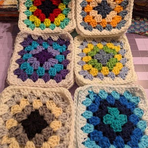 May include: Six colorful crocheted granny squares. Each square features a central design in various colors, including red, yellow, blue, and purple, surrounded by a white border. The squares are arranged on a purple surface.