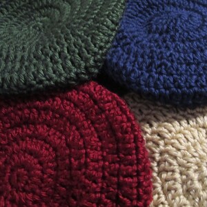 May include: Close-up of four crocheted hats in various colors: dark green, navy blue, burgundy, and beige. The hats are circular with a textured, spiral design. The image highlights the handmade quality and color variations.