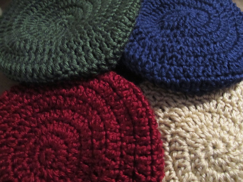 May include: Close-up of four crocheted hats in various colors: dark green, navy blue, burgundy, and beige. The hats are circular with a textured, spiral design. The image highlights the handmade quality and color variations.
