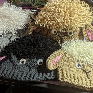May include: A collection of handmade crochet sheep hats in various colors, including white, tan, gray, and black. Each hat features a fluffy woolly top, googly eyes, and pink-lined ears. The hats are designed with a cute, whimsical aesthetic.