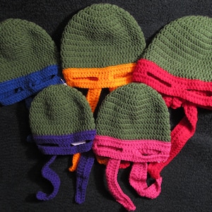 May include: Five crocheted hats in green, orange, red, purple, and pink. Each hat has a matching colored mask attached to the bottom. The hats are designed to look like the Teenage Mutant Ninja Turtles.