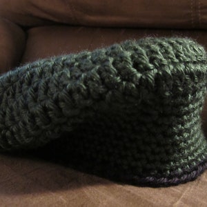 May include: A dark green, hand-crocheted hat with a rounded crown and a small, curved brim. The hat is made with a textured stitch, and the brim has a dark purple trim. The hat is likely for cold weather.