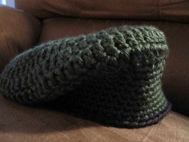 May include: A dark green, hand-crocheted hat with a rounded crown and a small, curved brim. The hat is made with a textured stitch, and the brim has a dark purple trim. The hat is likely for cold weather.