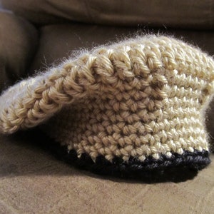 May include: A tan crocheted hat with a black brim. The hat is made of a thick yarn and has a textured appearance. The hat is sitting on a brown surface. The hat is a type of headwear.
