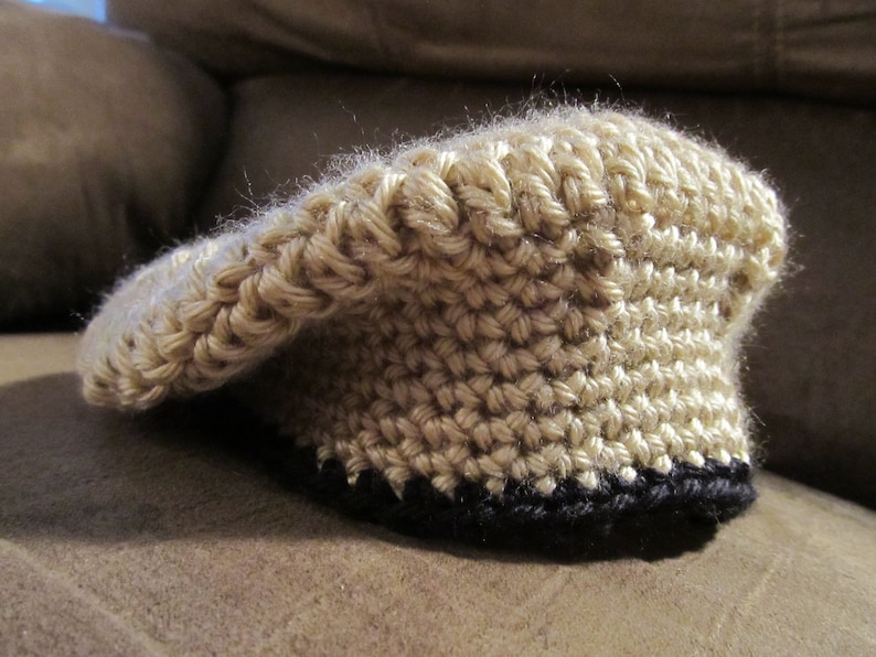 May include: A tan crocheted hat with a black brim. The hat is made of a thick yarn and has a textured appearance. The hat is sitting on a brown surface. The hat is a type of headwear.