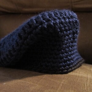 May include: A dark blue, hand-crocheted hat with a brim. The hat is made of a textured yarn, and the brim is a darker shade of blue. The hat is sitting on a brown surface.