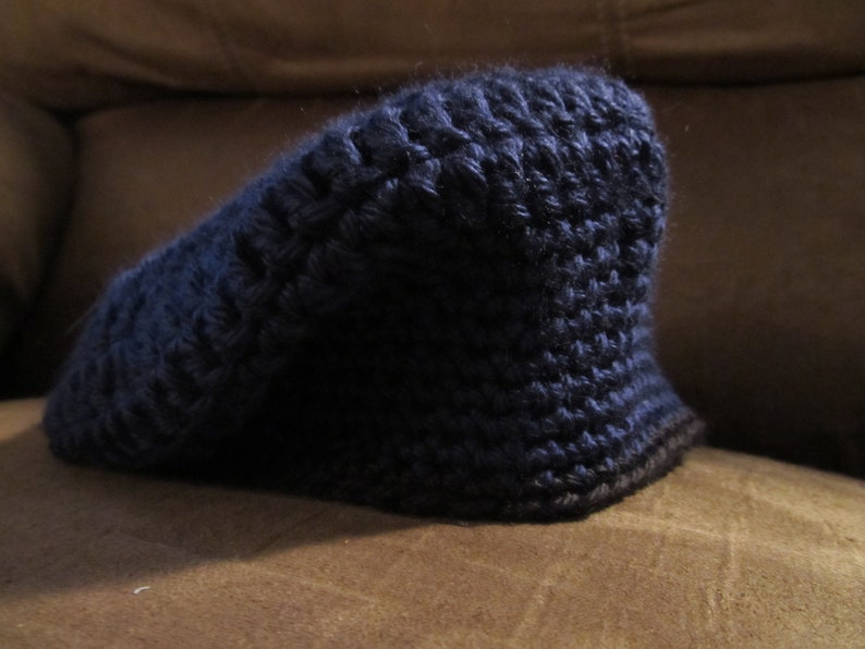 May include: A dark blue, hand-crocheted hat with a brim. The hat is made of a textured yarn, and the brim is a darker shade of blue. The hat is sitting on a brown surface.