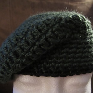 May include: A dark green, hand-crocheted beret hat. The hat is made of a textured yarn, with a slightly sloped crown and a rounded brim. The beret is displayed on a white mannequin head, against a brown background.