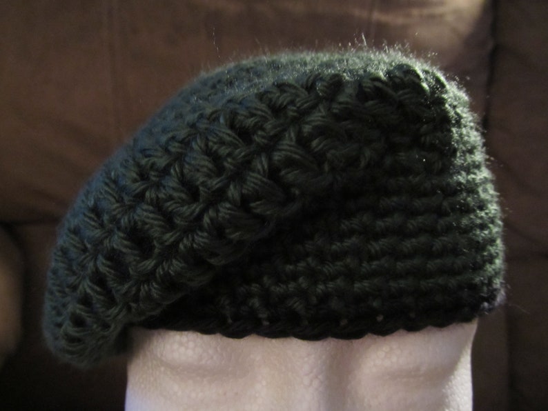 May include: A dark green, hand-crocheted beret hat. The hat is made of a textured yarn, with a slightly sloped crown and a rounded brim. The beret is displayed on a white mannequin head, against a brown background.
