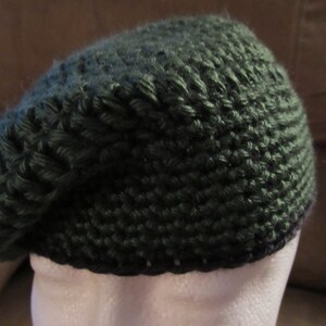 May include: A dark green knitted beret. The beret is made of a thick, textured yarn, and has a rounded crown and a brim that sits close to the head. The beret is displayed on a white mannequin head.