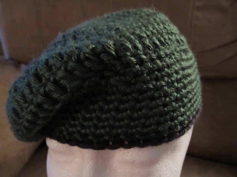 May include: A dark green knitted beret. The beret is made of a thick, textured yarn, and has a rounded crown and a brim that sits close to the head. The beret is displayed on a white mannequin head.