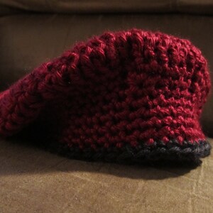 May include: A burgundy crochet hat with a black brim. The hat has a pointed top and is made of thick yarn. The hat is sitting on a brown surface.