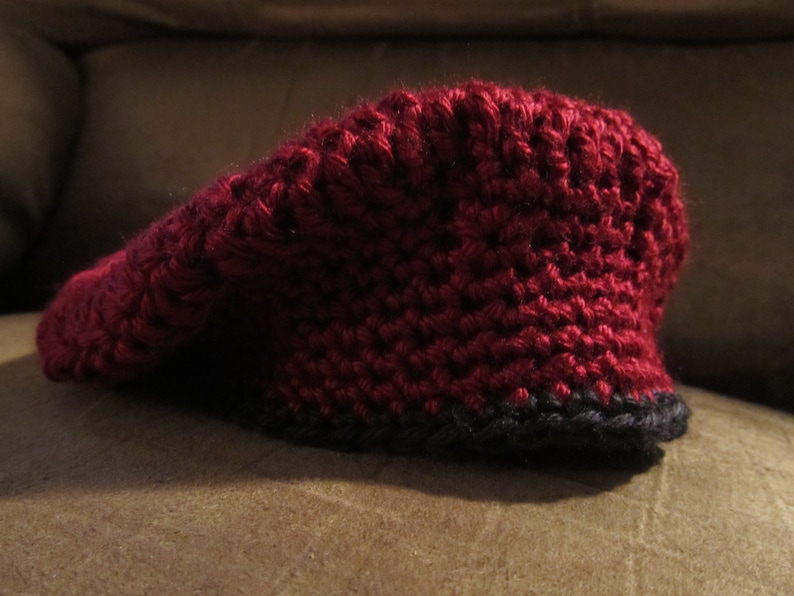 May include: A burgundy crochet hat with a black brim. The hat has a pointed top and is made of thick yarn. The hat is sitting on a brown surface.