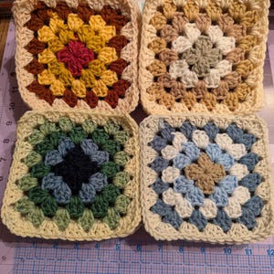 May include: Four handmade crochet granny squares in various color combinations. The squares feature a central floral design in shades of red, yellow, brown, green, blue, and cream. Each square is bordered with a cream-colored yarn.