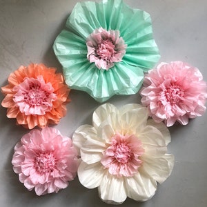 May include: Five tissue paper flowers in various colors, including peach, pink, and light green. The flowers are arranged on a gray surface.