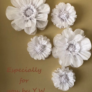 May include: Five white paper flowers of varying sizes are arranged on a beige background. The flowers are made of tissue paper and have a delicate, ruffled appearance. The text "Especially for you by Y.W." is written in brown cursive below the flowers.