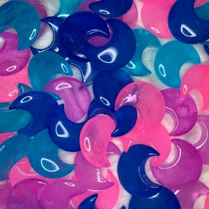 May include: A collection of crescent moon shaped crystal stones in shades of pink, blue, and purple. The stones are smooth and polished, with a translucent appearance.