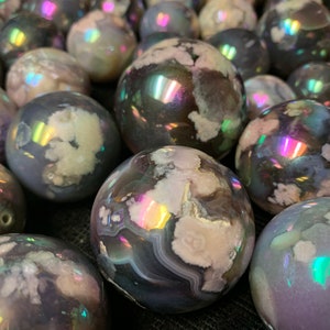 Puede incluir: Una colección de esferas de ágata iridiscente en tonos de morado, gris y blanco. Las esferas tienen una superficie lisa y pulida y están dispuestas en primer plano.