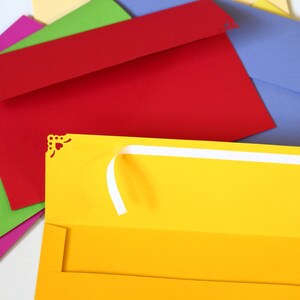May include: A stack of colorful envelopes with decorative edges. The top envelope is yellow and open, revealing a white ribbon.