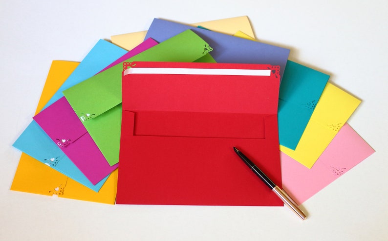 May include: A stack of colorful envelopes with decorative cutouts, a red envelope is open with a black pen on top of it.