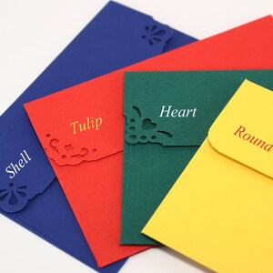 May include: Four different colored envelopes with decorative cutouts. The envelopes are blue, red, green, and yellow. The cutouts are shaped like a shell, tulip, heart, and circle. The envelopes are labeled with the names of the shapes.