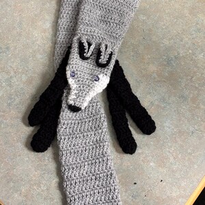 May include: A handmade crochet scarf in the shape of a fox. The scarf is primarily gray with black accents for the ears and legs, and a white tip on the tail. The fox face has purple eyes.