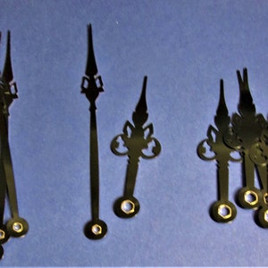 May include: Several black clock hands with decorative tips and gold-colored center holes are arranged on a blue surface. The hands vary in length and design, suitable for clock repair or crafting projects.