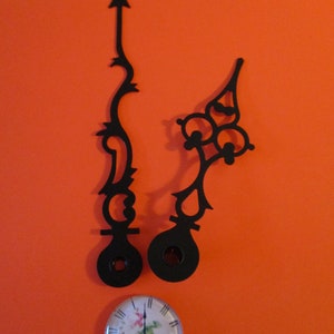 May include: Two black metal clock hands with intricate designs against an orange background. A small clock face with a floral design is in the bottom center.