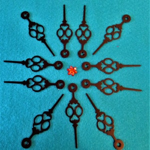 May include: A set of ten black clock hands with intricate designs. The hands are arranged in a circle on a turquoise surface.