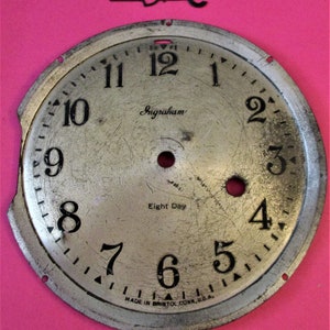 May include: A silver clock face with black numbers and hands. The clock face is marked "Ingraham" and "Eight Day" and "MADE IN BRISTOL CONN. U.S.A.".