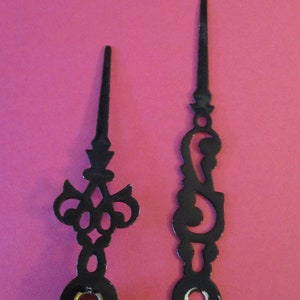 Könnte beinhalten: Zwei schwarze Uhrzeiger mit dekorativen Designs. Die Zeiger sind aus Metall und haben eine schwarze Oberfläche. Der größere Zeiger hat ein kreisförmiges Design am Ende, während der kleinere Zeiger ein komplizierteres Design hat.