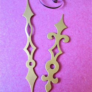May include: Two gold-coloured clock hands with intricate designs. The hand on the left is a simple, straight design with a diamond shape at the end. The hand on the right has a more ornate design with a fleur-de-lis at the end.