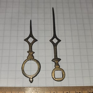 May include: Two gold-colored clock hands with a diamond-shaped design at the top. The hands are about 6 inches and 8 inches long. The hands are on a white gridded surface with a wooden ruler.